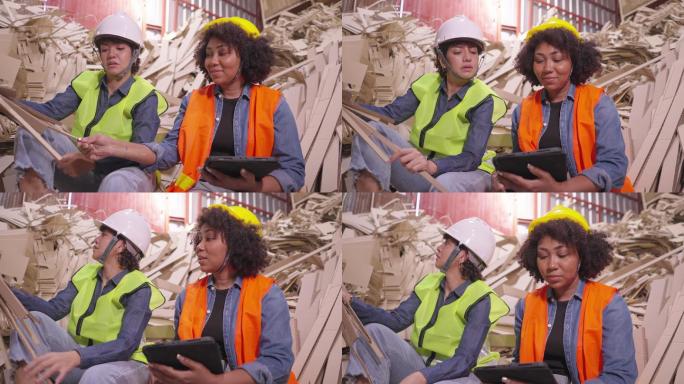 拉丁和非洲女性的可持续生活方式纸箱厂里，穿着安全背心和头盔的回收工人坐在一堆废纸板上，用数字平板电脑