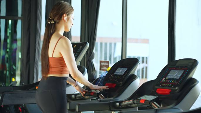 亚洲美丽的年轻运动女性，在跑步机上跑步，喝矿泉水，在健身房的智能手表上查看运动数据。