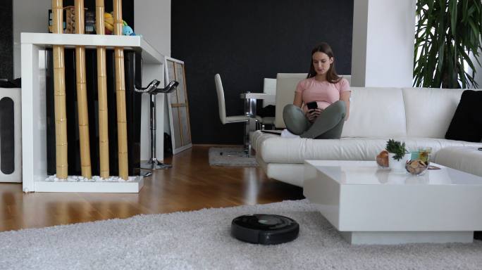 孕妇正在沙发上休息和享受。她通过自己的手机控制机器人吸尘器来清洁地毯