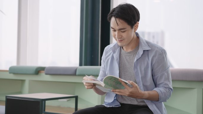 亚洲学生男子看书，坐在大学图书馆的办公空间里。一个黑发男子穿着蓝色衬衫，专注、专注、快乐。