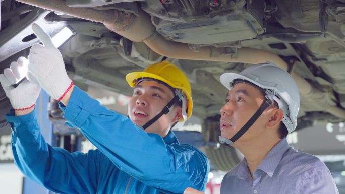 两名亚洲人在汽车修理厂戴头盔工作。车辆服务经理团队男性工人查看车辆状况，检查和维护，以便在车间维修发