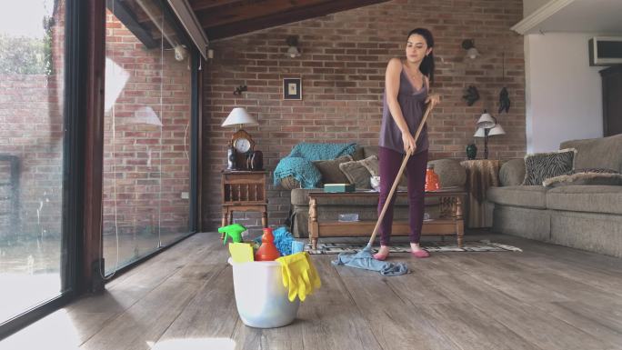 一名西班牙裔年轻女子在客厅用拖把和装有清洁用品的塑料桶清洁硬木地板