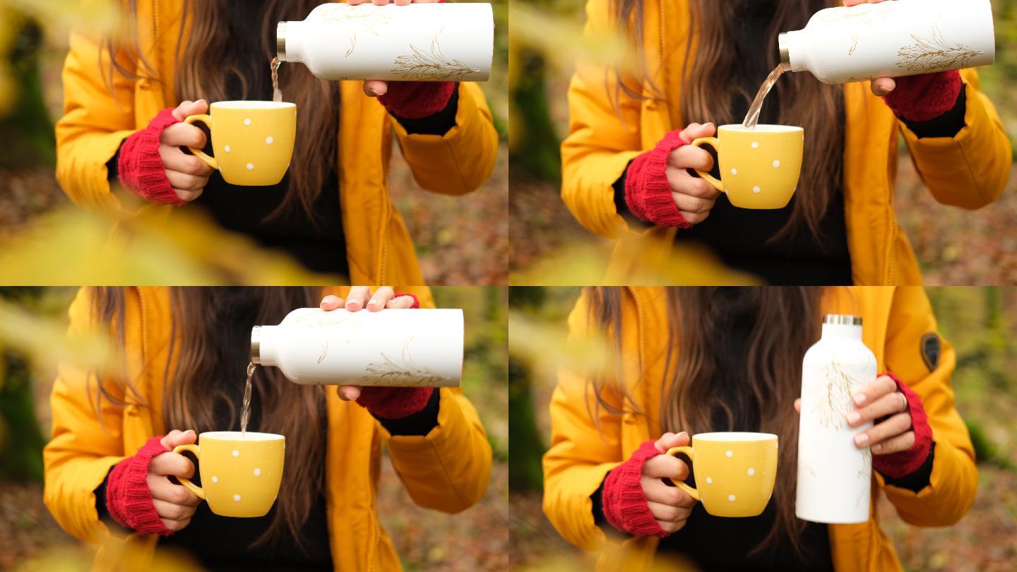森林里，一个穿着黄色派克大衣的女人从保温瓶里倒茶。