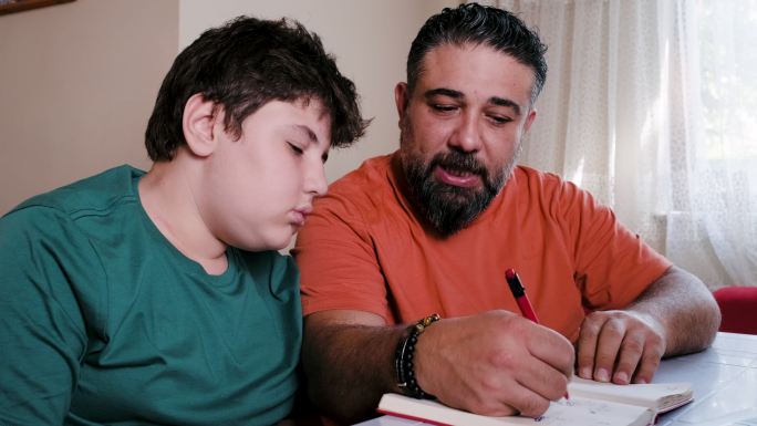 愤怒的父亲白天坐在笔记本电脑前的桌子旁，帮助压力重重的十几岁儿子做家庭作业