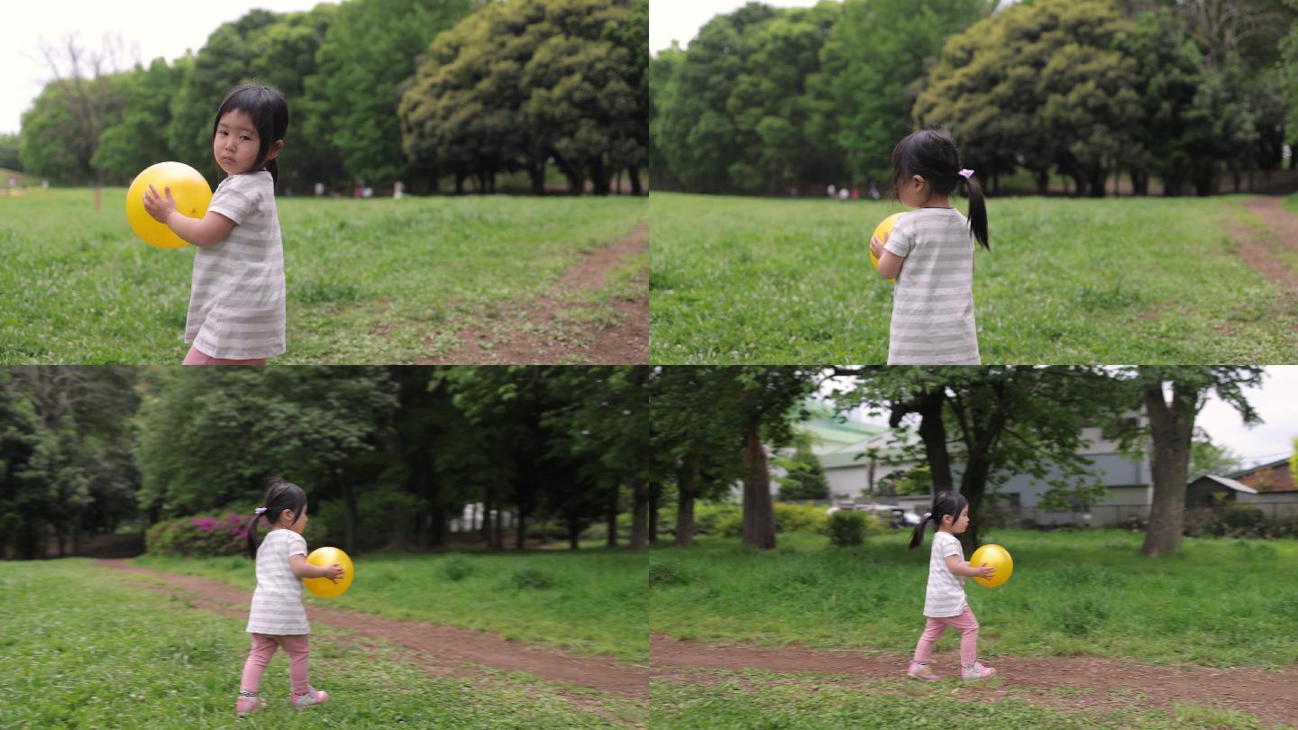 小女孩手里拿着黄色的大球，走在绿色的草地上