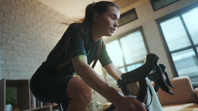 亚洲女自行车手。她正在屋里锻炼。通过在教练机上骑自行车和玩在线自行车游戏