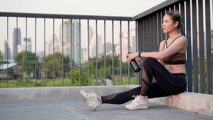 亚洲美丽的中年女性，穿着运动装，肌肉发达，坐在人行桥上休息，手上流汗，饮用城市公园里可重复使用的水瓶