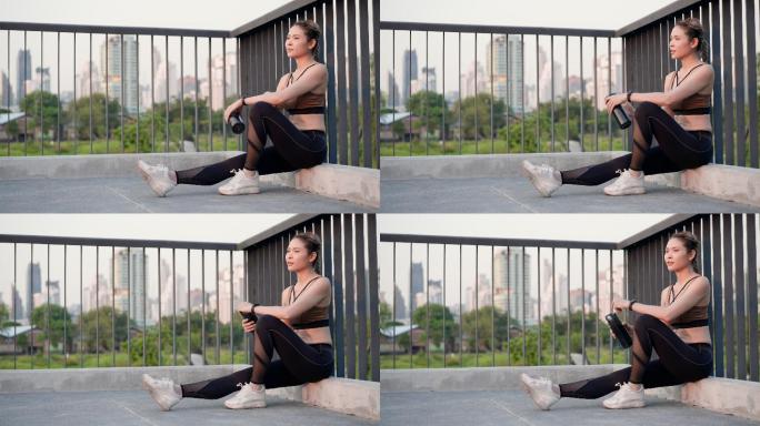 亚洲美丽的中年女性，穿着运动装，肌肉发达，坐在人行桥上休息，手上流汗，饮用城市公园里可重复使用的水瓶
