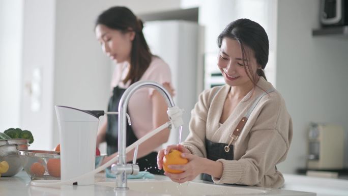 周末，亚裔中国女性在家中用笔记本电脑在厨房柜台用自来水清洗水槽中的橙色水果