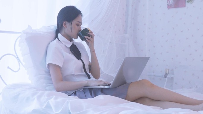 一名女性大学生在床用笔记本上网课喝水
