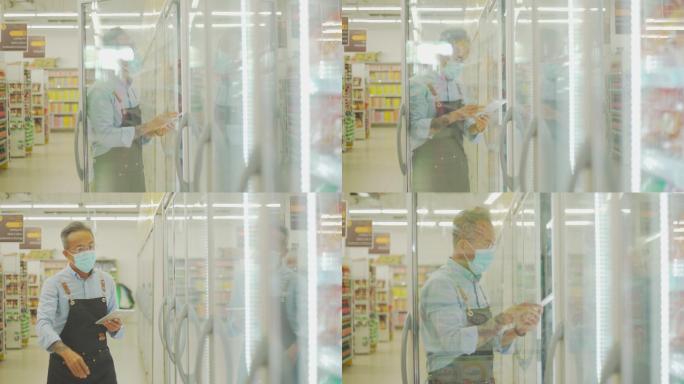 一名戴着口罩的亚裔中国老人在超市冷藏区用数码平板电脑清点冷冻食品