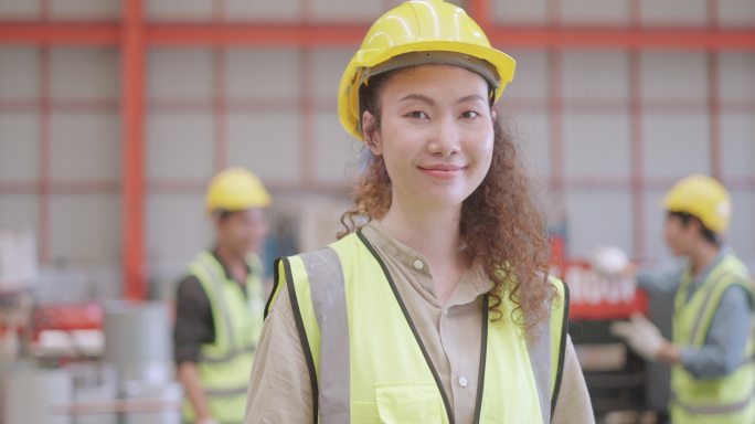 专业工业女性工程师或技师的肖像，有自信和领导力，穿戴个人防护装备，站在制造工厂，微笑着看着相机。