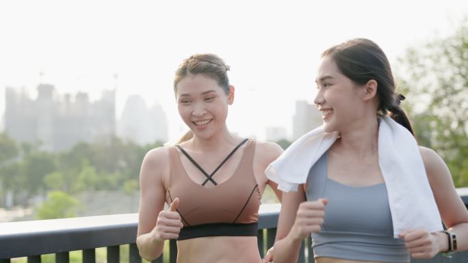 正面图：两位亚洲美丽的中年女性朋友穿着运动装，肌肉发达，早上一起在市中心附近的公园花园的人行桥上积极