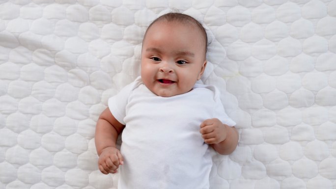 特写镜头：一个穿着白色婴儿服的无辜黑人新生儿躺在柔软的床上，表达婴儿的语言，上下抬起腿，微笑着看着相