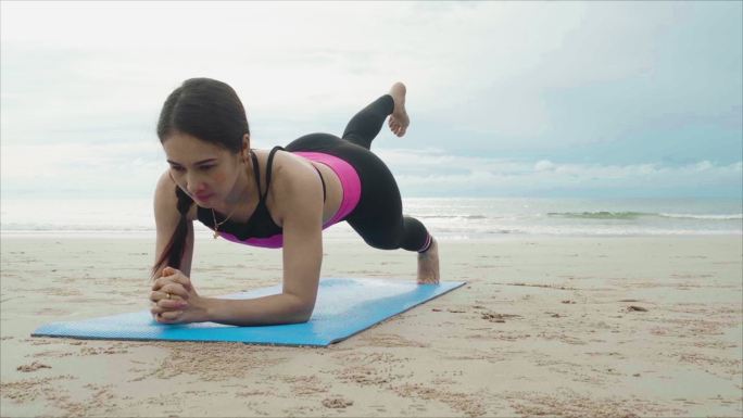 在瑜伽垫上做瑜伽板姿势的女人。早上，亚洲女性在海天多云的海滩上穿着运动服。