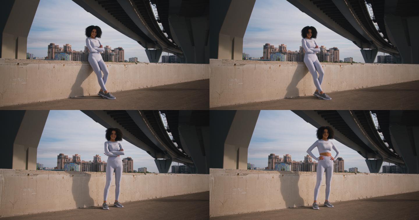 一位年轻美丽的跑步者在现代混凝土桥下的路堤上摆姿势，背景是城市景观