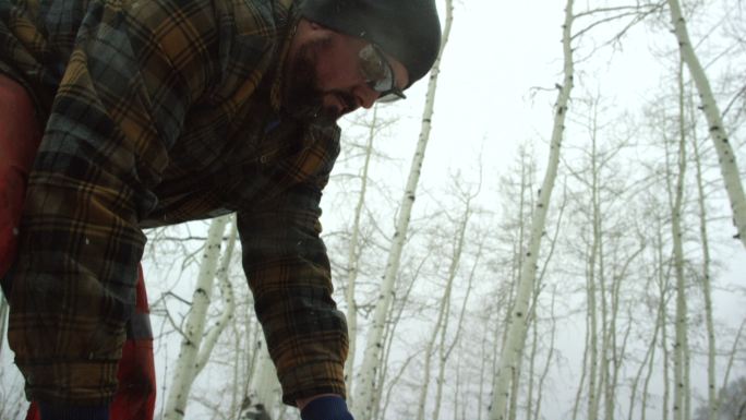 一名30多岁的白人男子在大雪纷飞的冬日，在山上用电锯切割一根白杨木原木