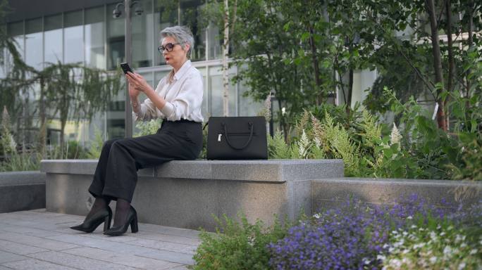 一位头发灰白、身穿白衬衫的漂亮女商人坐在商务中心附近的一张单色长椅上，用手机在线购物