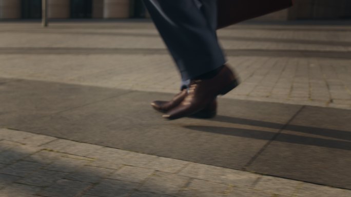 一个商人的脚穿着棕色皮鞋，从画面的右侧到左侧在铺好的街道上走过，
