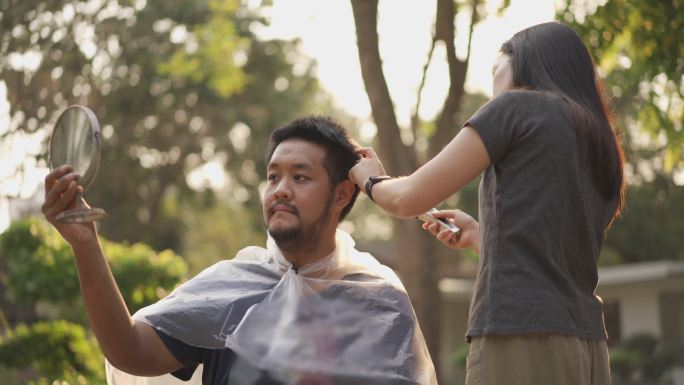 一名亚洲男子拿着镜子在家中被其夫妇在新冠病毒疫情中剪头发