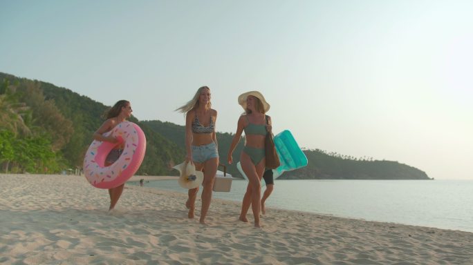 一群年轻的多民族朋友，他们一边散步，一边互相帮助，一边提着橡胶圈，塑料冷却器，一边在沙滩上享受日落的