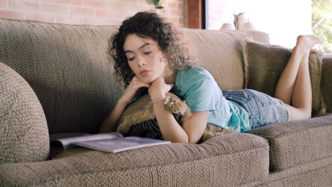 拉美裔卷发年轻女子在沙发上放松时看杂志