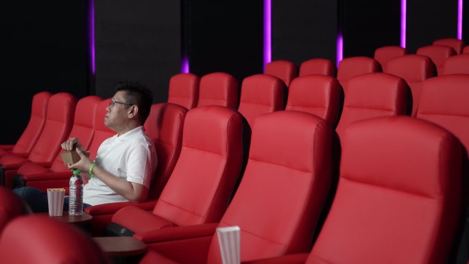 一名亚裔中国男子坐在空荡荡的电影院红色座位上，一边吃一边看电影。