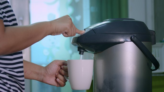 手上的特写镜头是按下电热锅上的按钮，将热水倒入陶瓷玻璃中。在厨房里，一位女士推着电热壶上的热水按钮喝