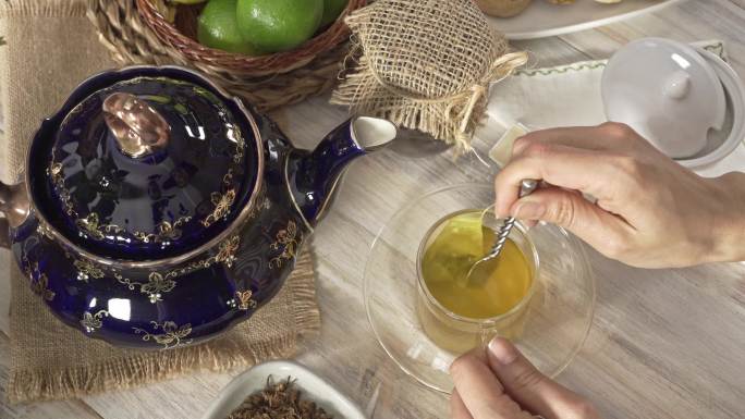 白色乡村桌子上，一位女士双手将茶包搅拌到热茶甘菊输液杯中的高角度特写镜头
