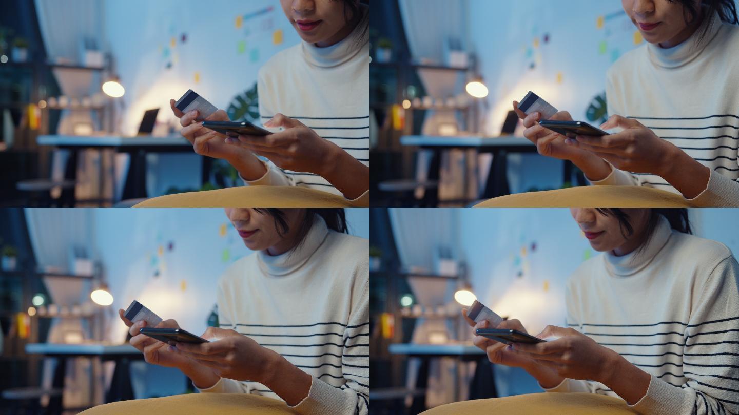 年轻的亚洲女性晚上坐在家里的沙发上购物时，一边用手机一边用信用卡进行在线交易。