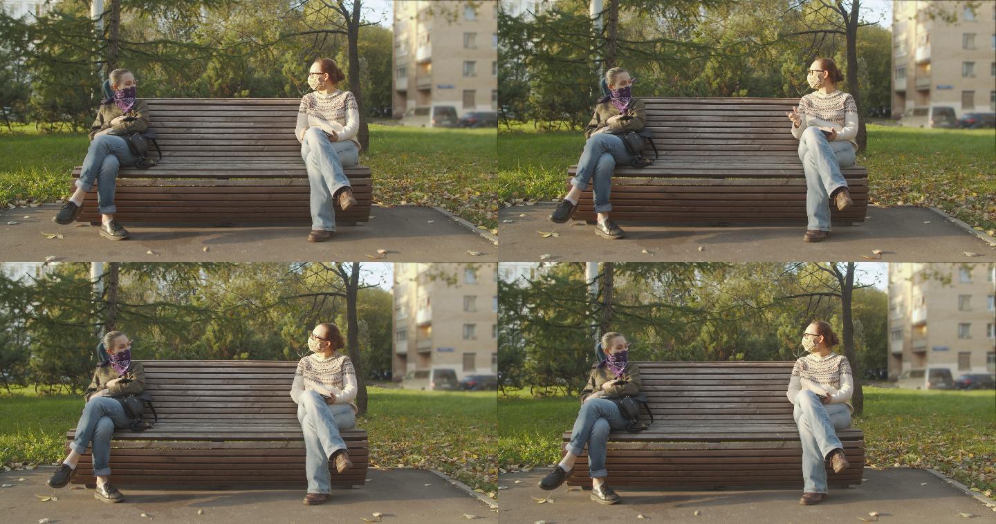 两名女性戴着一次性防护面罩或手帕面罩，坐在长凳上，保持社交距离