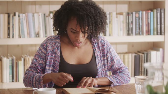 一位非洲裔女性在客厅里读着迷人的书。情节扭曲的情绪反应-看着相机
