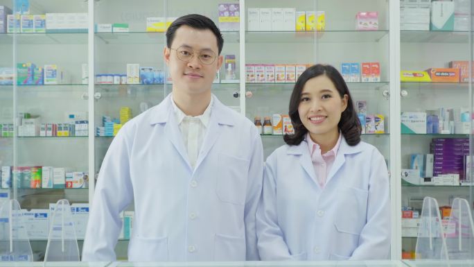 药房背景中的亚洲药剂师肖像。药剂师微笑着看着药房里的摄像机，用药盘数药丸并分发给患者。医疗保健和医疗