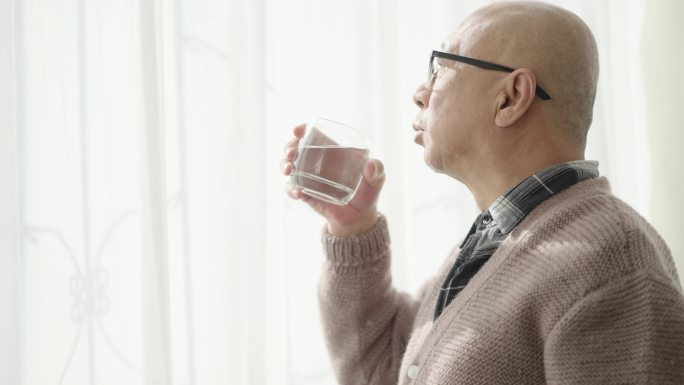 一位中国老人站在窗边，一只手拿着一杯水吃药