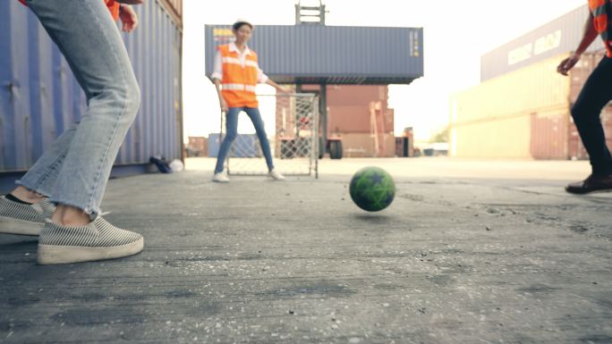 在货柜场的院子里，一名穿着反光衣服和裤子的工程督察工头充当足球守门员，试图拦截一名同事踢出的足球。