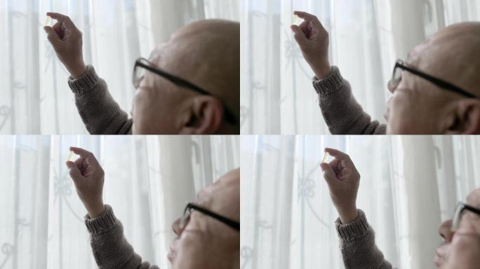 一位中国老人坐在窗边，手里拿着一个胶囊，戴着阅读眼镜看着