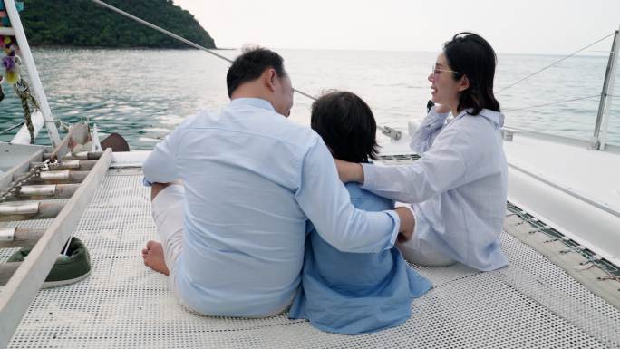 后视图：一家三口人，父亲、母亲和儿子坐在一艘豪华游艇上的蹦床上，一起在海上聊天。儿子坐在爸爸妈妈中间