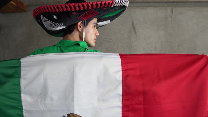 一个十几岁的拉丁男孩的肖像，他举着墨西哥国旗，头戴一顶罩衫
