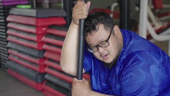 超重男子在健身房锻炼后，气喘吁吁地坐在健身机上，脸上非常疲倦。