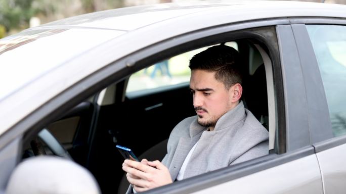 坐在车里用手机打在线信息的年轻人，侧视图。微笑的商人在汽车里使用手机。