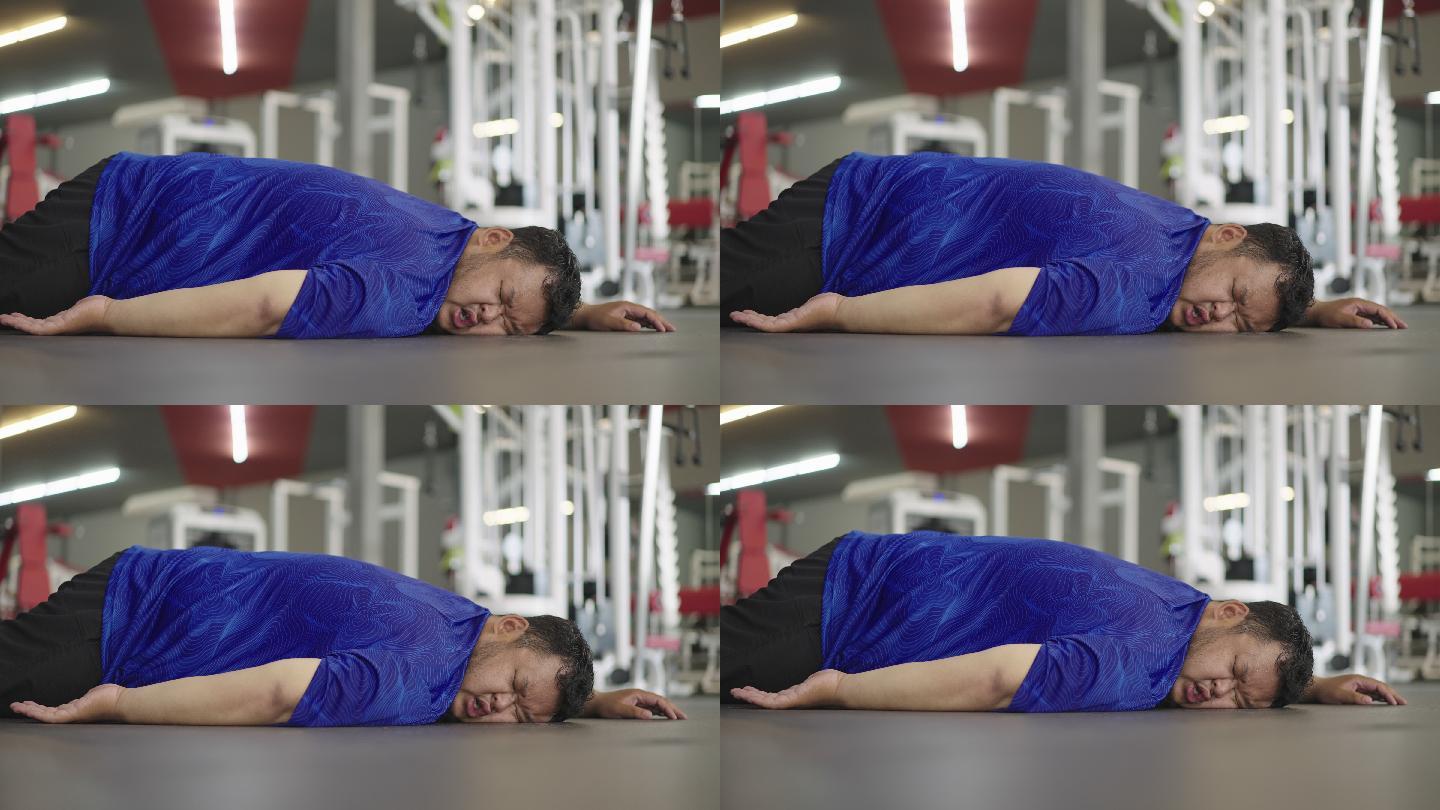 一个非常胖的家伙趴在地上喘着气，一边在健身房健身休息。