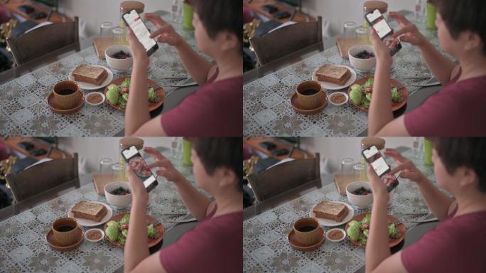 回头看中国亚裔女性早上在餐桌上吃健康的沙拉早餐时，用智能手机上网滚动社交媒体