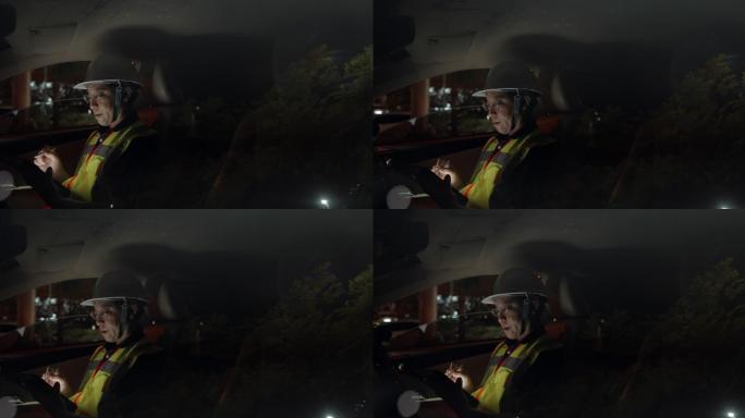 亚洲土木工程师男子戴安全帽，穿安全背心在工地上夜班。工头坐在车上，在数字平板上检查结构的进度，并在城