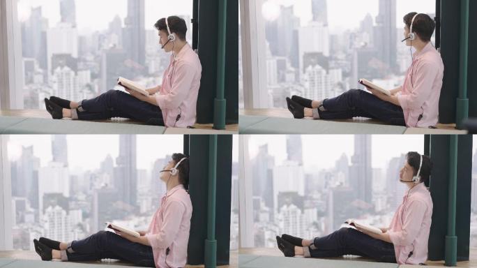 在高楼校园图书馆，帅气的亚洲大学生男子坐在窗户附近，可以看到很好的景色，放松时间戴着耳机听音乐，一边
