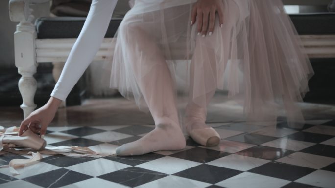 美丽的年轻芭蕾舞演员在工作室的地板上系着鞋子，鞋子上系着丝带。