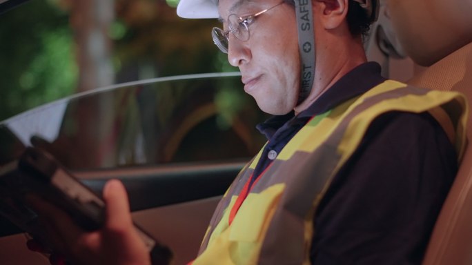 亚洲工程师戴安全帽，穿安全背心在工地上夜班。工业工头工作到很晚，坐在车上，用数字平板电脑检查计划，监
