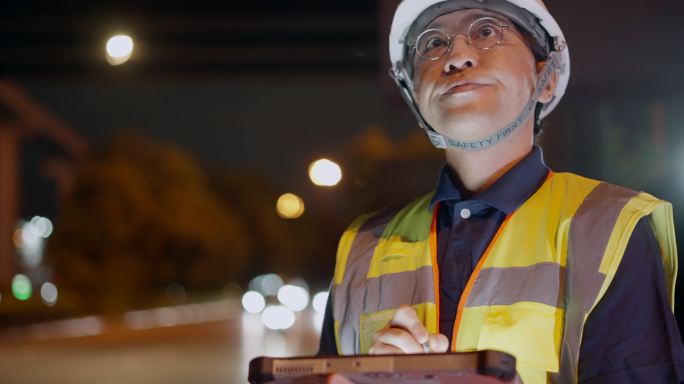 亚洲男性土木工程师或建筑师在互通式高速公路施工现场戴安全帽、穿安全背心。值夜班时，工头在数字平板上行