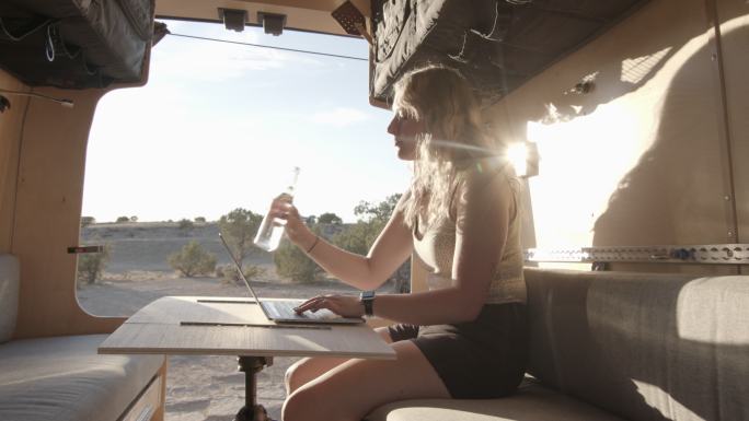 科罗拉多州弗鲁塔附近，一位金发碧眼的千禧一代年轻女性开着定制露营车，用笔记本电脑从瓶子里喝水
