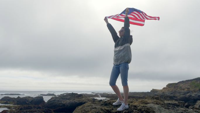 太平洋野生海岸岩石上挂着美国国旗的女人。加利福尼亚