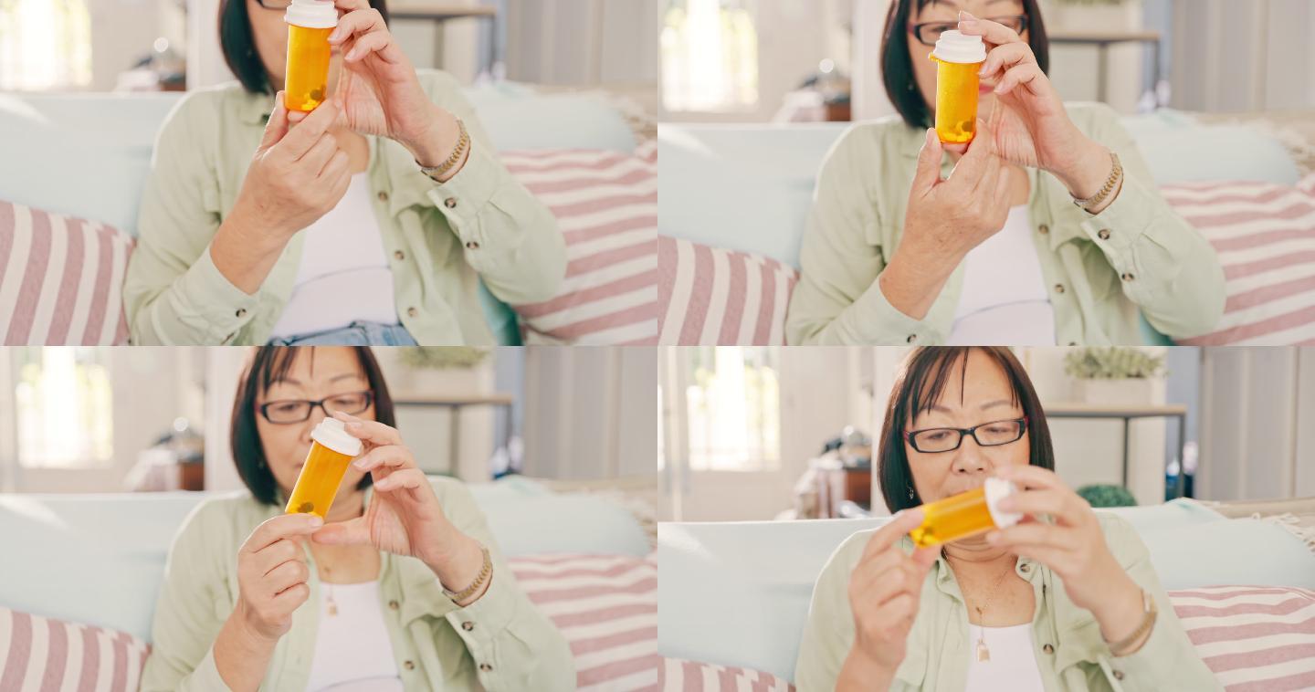 一位亚洲老妇人独自坐在家里，看着一瓶药丸。生病的亚洲老妇人在家的客厅里拿着药